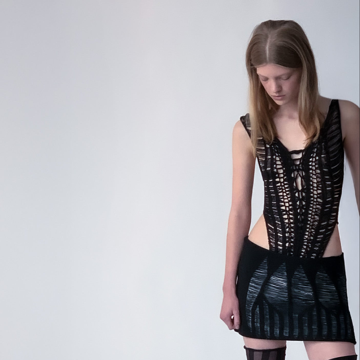 Person wearing a black knitted bodice and a white leather and black knit mini skirt on light grey background 