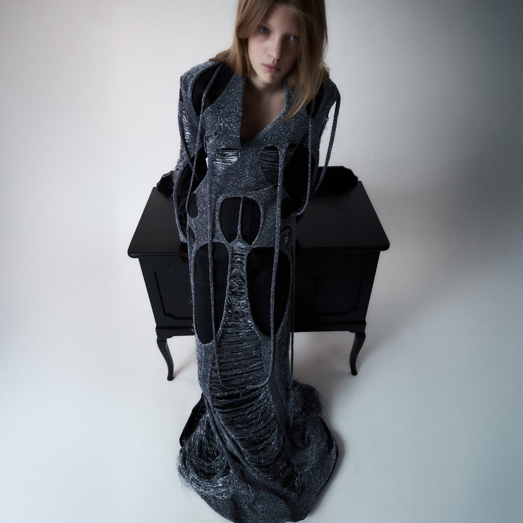 Woman wearing a long, dark gray dress layered with metallic knit standing next to a black desk.