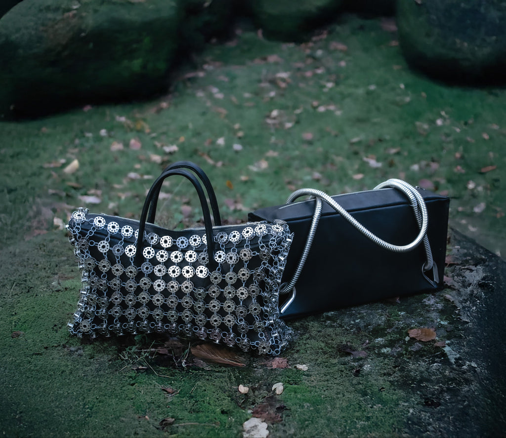 Two handbags, one with metal bobbins embellishment and the other black leather with metal handles on a grassy surface.
