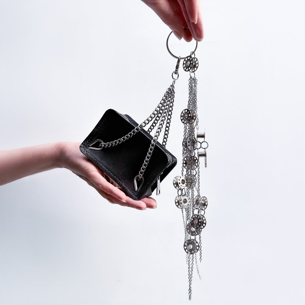  mini black leather bag with metal chain handles as a bag's charm  with a metal-bobbins and metal-chains composition bag's charm on a light gray background